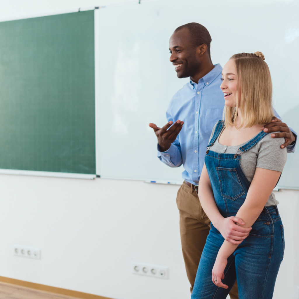 Male teacher and fmeal high school student standing closely in front of chalk and whiteboard. The student introduced themself and now the teacher is introducing her to the class. 