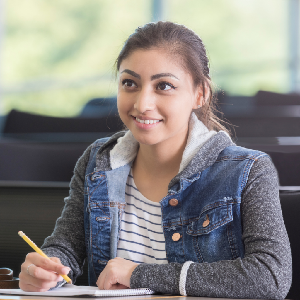 8th or Ninth grade femaile student, smiling, pencil in hand, at her desk in school and looking up. 